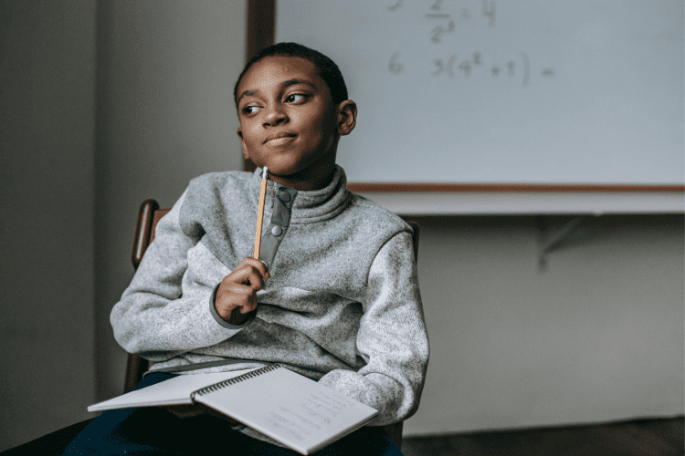 a boy in the classroom , using a pencil pointing on his chin , open book